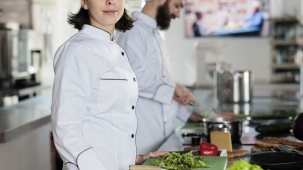 Portrait of beautiful happy sous chef standing in restaurant professional kitchen, wearing cooking uniform while smiling at camera. Young adult food industry worker preparing vegetables for meal.