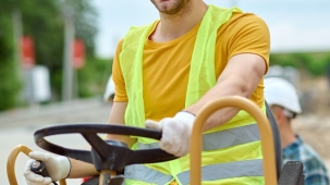 Optimism. Young adult smiling man in protective helmet yellow tshirt and bright vest sitting behind wheel of special construction equipment looking at camera outdoors