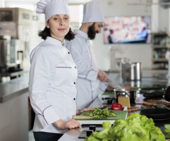 Portrait of beautiful happy sous chef standing in restaurant professional kitchen, wearing cooking uniform while smiling at camera. Young adult food industry worker preparing vegetables for meal.